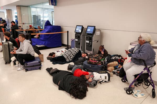Stranded travelers waiting at an airport terminal with flight information screens showing cancellations.